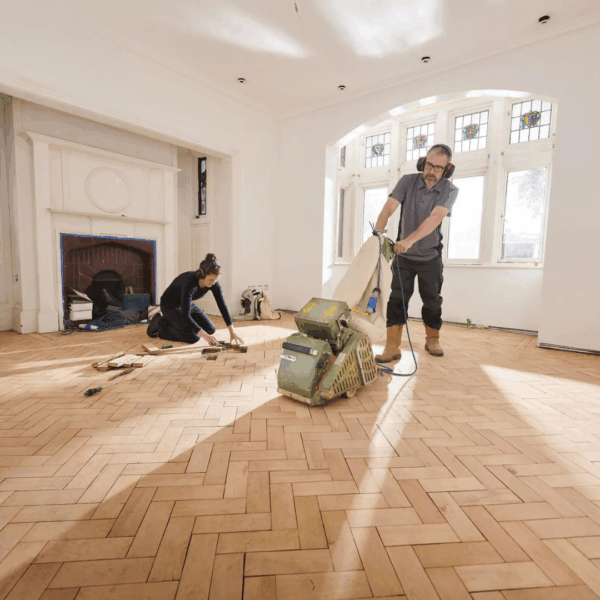 Two workers renovating a room with herringbone hardwood floors; one is sanding while the other is working on the ground.